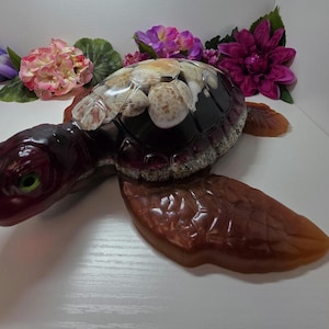 May include: A sea turtle figurine crafted from dark brown resin with a glossy finish. The shell is embedded with various natural seashells, and the turtle features bright green eyes. The flippers and head have a translucent, amber-toned appearance.