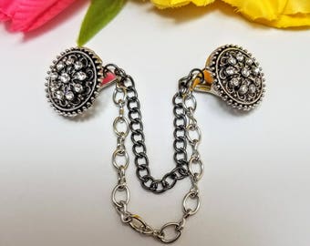 May include: A pair of silver tone decorative chain clasps with clear rhinestone accents. The clasps are connected by a silver and black chain.