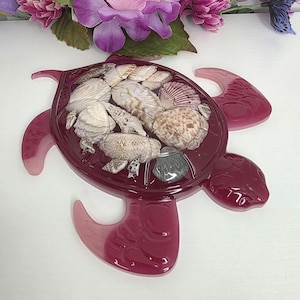 May include: A turtle-shaped decorative tray made of translucent deep magenta resin. The shell area is embedded with a variety of natural seashells in shades of white, tan, and grey, creating a textured mosaic pattern within the smooth resin surface.
