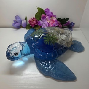 May include: A blue sea turtle figurine crafted from translucent resin. The shell features an embedded miniature scene with sand, small white seashells, and a tiny green palm tree. The turtle has a smooth, glossy finish and detailed scales.