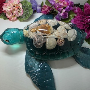 May include: A decorative sea turtle figurine crafted from translucent teal resin. The shell is embedded with a variety of natural seashells in shades of white, cream, and brown, creating a textured, multi-dimensional appearance.