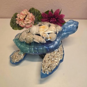 May include: A decorative sea turtle figurine crafted from blue and teal resin. The shell is filled with various white and tan seashells, while the flippers are embedded with small crushed white shells, creating a textured, ocean-inspired appearance.