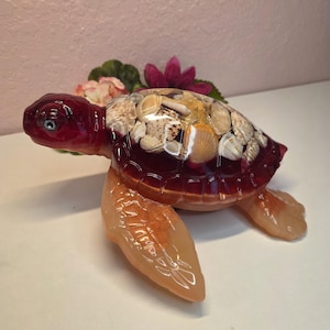 May include: A decorative sea turtle figurine crafted from translucent resin. The head and limbs are a warm amber tone, while the shell is a deep burgundy color filled with an assortment of embedded natural seashells and a small starfish.
