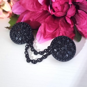 Könnte beinhalten: Ein Paar schwarze, runde Zierknöpfe, die durch eine Kette verbunden sind. Jeder Knopf hat ein detailliertes, erhabenes Blumenmuster und einen Perlenrand. Die Knöpfe sind vor einem Hintergrund aus rosa und magenta Blüten platziert.