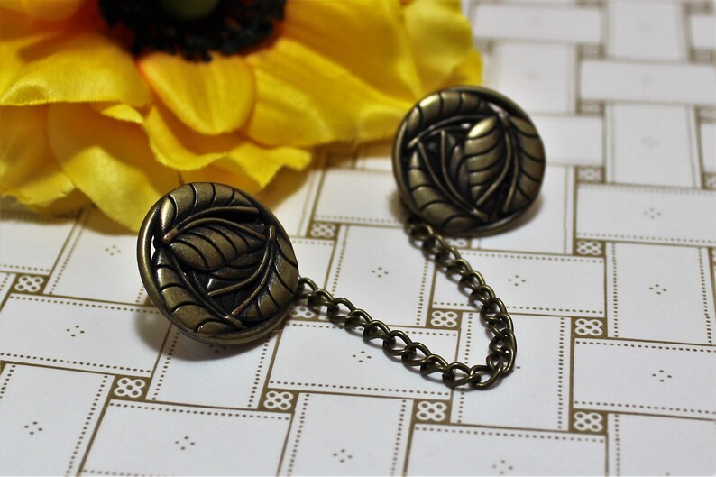 Puede incluir: Un par de broches de metal dorado antiguo con dise&ntilde;o de hojas conectadas por una cadena.
