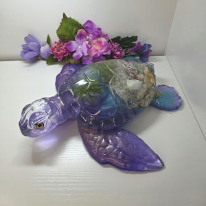 May include: A translucent purple resin sea turtle figurine featuring a clear shell filled with embedded sand, small seashells, and green moss-like elements. The turtle has a smooth, glossy finish and is positioned with its flippers extended.