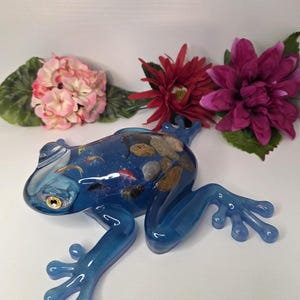 May include: A blue resin frog figurine with embedded stones and small fish. The frog has a glossy finish and is posed with its legs extended. Pink and red flowers are arranged near the frog.