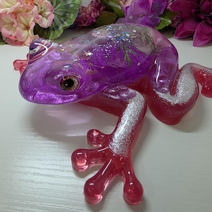May include: A translucent resin frog figurine featuring a vibrant purple head and back embedded with glitter and dried botanical elements. The limbs are a deep pink hue with silver glitter accents on the legs and a realistic brown eye detail.