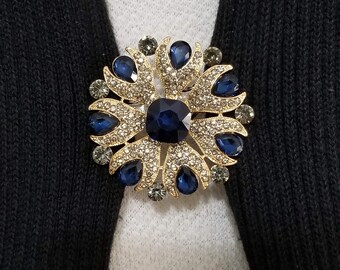 May include: A gold-toned brooch with a blue gemstone centre and smaller clear gemstones around the edges. The brooch is shaped like a flower with pointed petals.