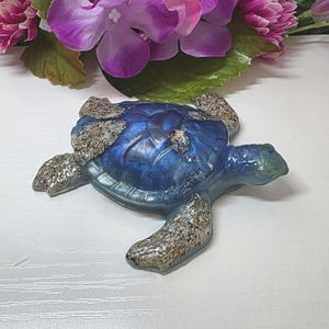 May include: A sea turtle figurine featuring a deep blue, pearlescent shell and head, with textured, sandy-colored flippers that appear to contain small stone or shell fragments embedded within the resin material.