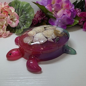 May include: A turtle-shaped trinket box made of translucent resin with a glittery magenta and purple gradient finish. The shell features an embedded collection of various natural seashells and a small black and white striped insect figurine.