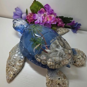 May include: A decorative sea turtle figurine featuring a translucent blue resin shell filled with miniature palm trees and white seashells. The head, flippers, and tail are textured with embedded sand and small pebbles, creating a realistic coastal appearance.