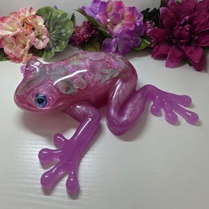 May include: A translucent purple resin frog figurine featuring embedded floral patterns, butterfly motifs, and shimmering glitter throughout its body. The frog has prominent, realistic blue eyes and a smooth, glossy finish, capturing a detailed amphibian form.