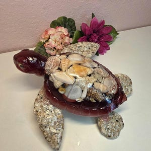 May include: A decorative turtle figurine with a dark red shell and head, adorned with seashells. The turtle's shell is filled with various seashells. Pink and purple flowers are placed on the turtle's back.
