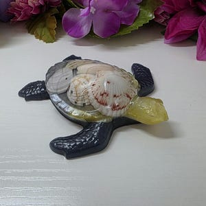 May include: A decorative sea turtle figurine featuring a dark gray textured body and a translucent shell filled with various small white and brown seashells. The turtle has a pale yellow head and flippers that extend outward from the central shell.