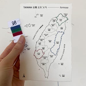 May include: Hand-stitched map of Taiwan on textured paper, with embroidered outlines of cities in blue, green, and red thread. The map includes text in English and Chinese, with a needle and thread spool in the upper left corner.