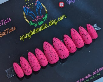May include: A set of ten pink press-on nails with black speckles. The nails are arranged in a row on a black surface. The text "Spicy Bite Nails" is visible in the upper right corner of the image.