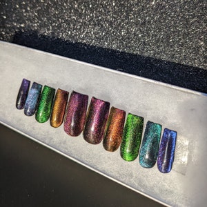 Op de afbeelding: Een set van tien iriserende, zelfklevende nagels in verschillende tinten groen, blauw en paars. De nagels zijn in een rij op een wit oppervlak gerangschikt.