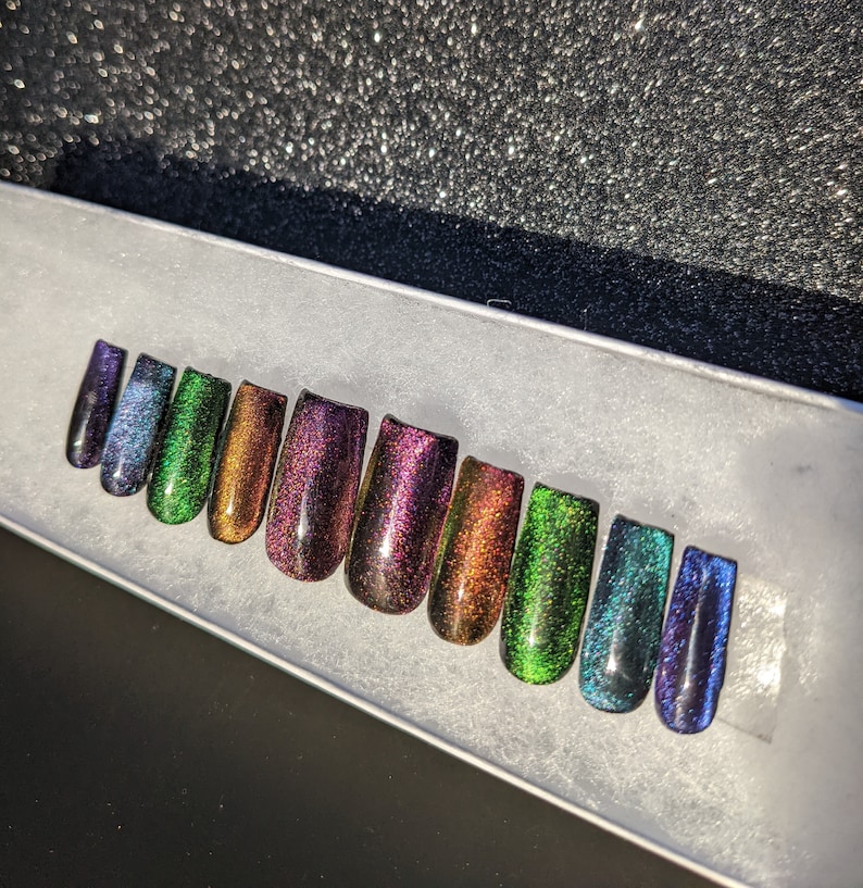 Op de afbeelding: Een set van tien iriserende, zelfklevende nagels in verschillende tinten groen, blauw en paars. De nagels zijn in een rij op een wit oppervlak gerangschikt.