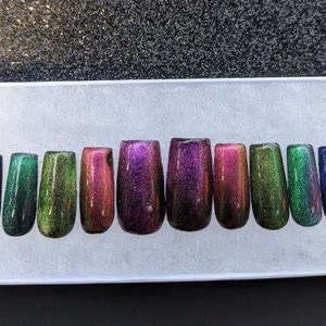 Op de afbeelding: Tien iriserende press-on nagels in verschillende kleuren, waaronder groen, paars en rood. De nagels zijn in een rij op een wit oppervlak gerangschikt.