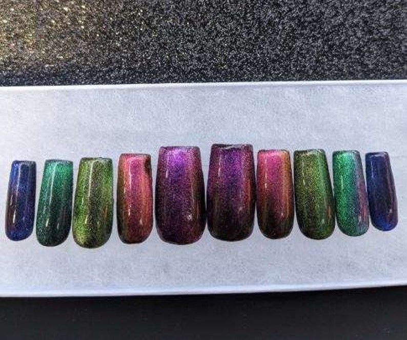 Op de afbeelding: Tien iriserende press-on nagels in verschillende kleuren, waaronder groen, paars en rood. De nagels zijn in een rij op een wit oppervlak gerangschikt.