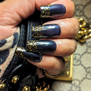 Puede incluir: Esmalte de uñas azul marino oscuro con detalles dorados con estampado de leopardo. Las uñas largas y cuadradas están adornadas con un diseño dorado en las puntas. Las uñas se sostienen cerca de un bolso de cuero negro con herrajes dorados.