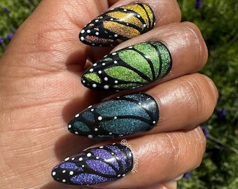 May include: Almond-shaped press-on nails with a butterfly wing design. The nails feature a black base with colorful glitter and white polka dots. The colors include gold, green, teal, and purple, mimicking butterfly wings.