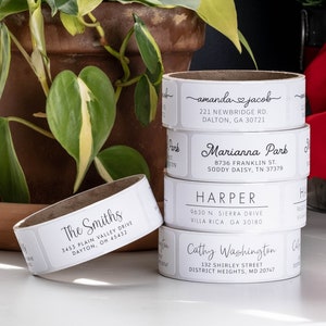 May include: A stack of white address labels with black text. The labels are for different people and addresses. The labels are on a roll and are ready to be used.