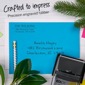 May include: A blue envelope with address information and a rubber stamp. The text "Crafted to impress" and "Precision engraved rubber" are at the top. The stamp is black with a white label that says "COLOP". A pencil and greenery are also in the image.