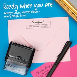 May include: A black and clear self-inking stamp with the text "COLOP printer C 40" next to a pink envelope with the address "Sanford" and a black pen. The background is blue with the text "Ready when you are!"