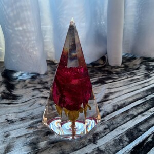 Op de afbeelding: Een heldere glazen piramide met een geconserveerde rode roos erin. De piramide staat op een grijs en zwart stoffen oppervlak.