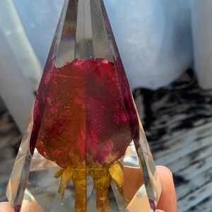 Op de afbeelding: Een heldere glazen piramide met een gedroogde rode roos erin. De roos is geconserveerd in hars en heeft een gouden steel.