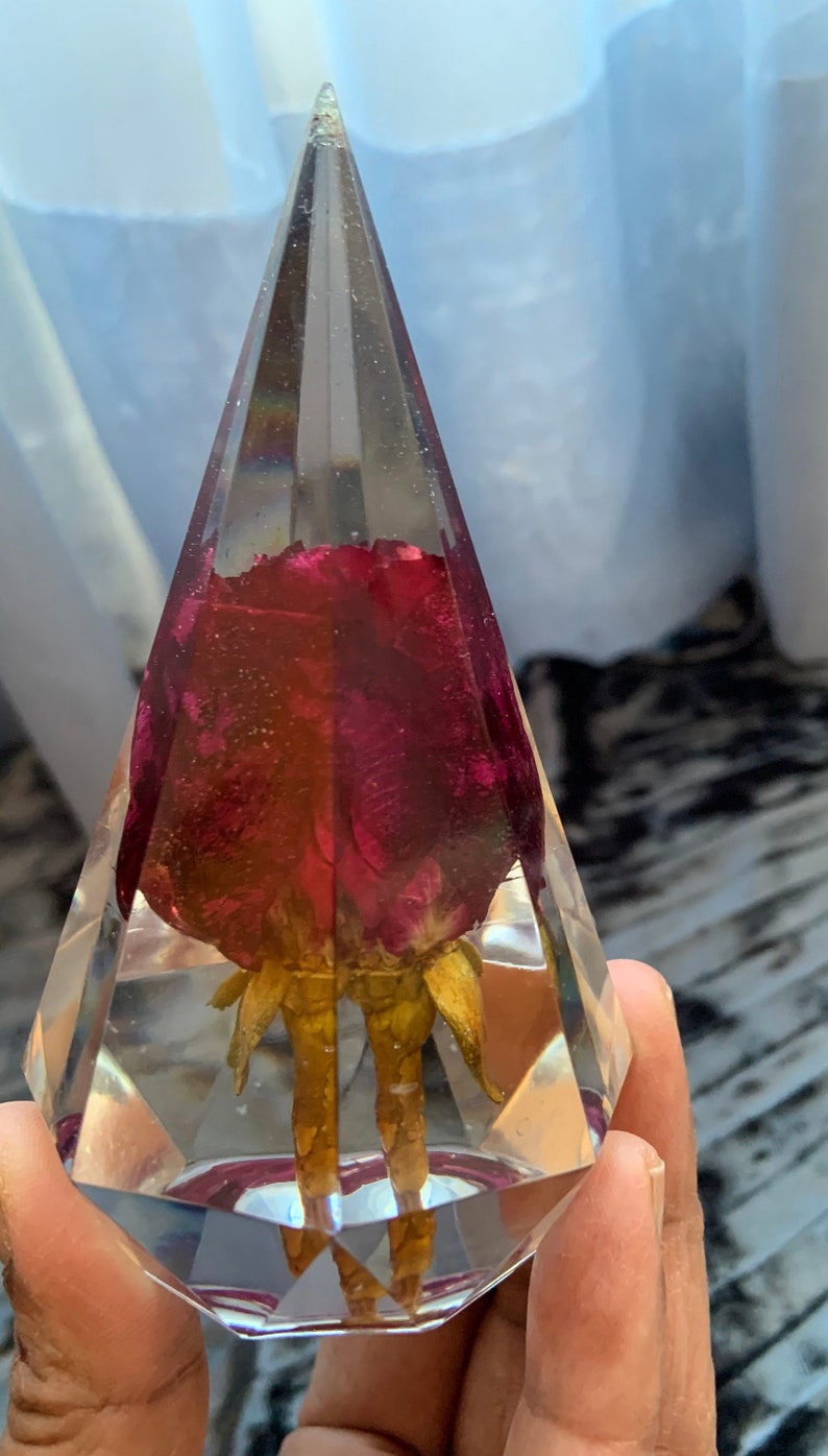 Op de afbeelding: Een heldere glazen piramide met een gedroogde rode roos erin. De roos is geconserveerd in hars en heeft een gouden steel.