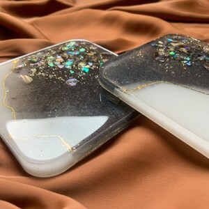 May include: Two square black and white resin coasters with gold accents and iridescent glitter. The coasters are on a brown surface.