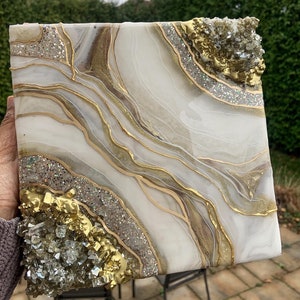 Könnte beinhalten: Abstrakte Harz-Kunst in Weiß und Gold mit Glitzer- und Blattgold-Akzenten. Die Kunst zeigt ein geodenartiges Design mit goldenen und klaren Kristallen.