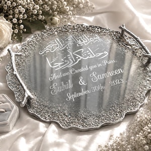 May include: A silver decorative tray with a textured edge and branch-style handles. The tray features engraved Arabic and English text, including names and a date. A ring box and white flowers are also visible.