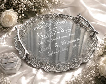 Könnte beinhalten: Ein silberfarbenes Dekotablett mit strukturiertem Rand und astähnlichen Griffen. Das Tablett hat gravierten Text in Arabisch und Englisch, einschließlich Namen und einem Datum. Eine Ringbox und weiße Blumen sind ebenfalls vorhanden.