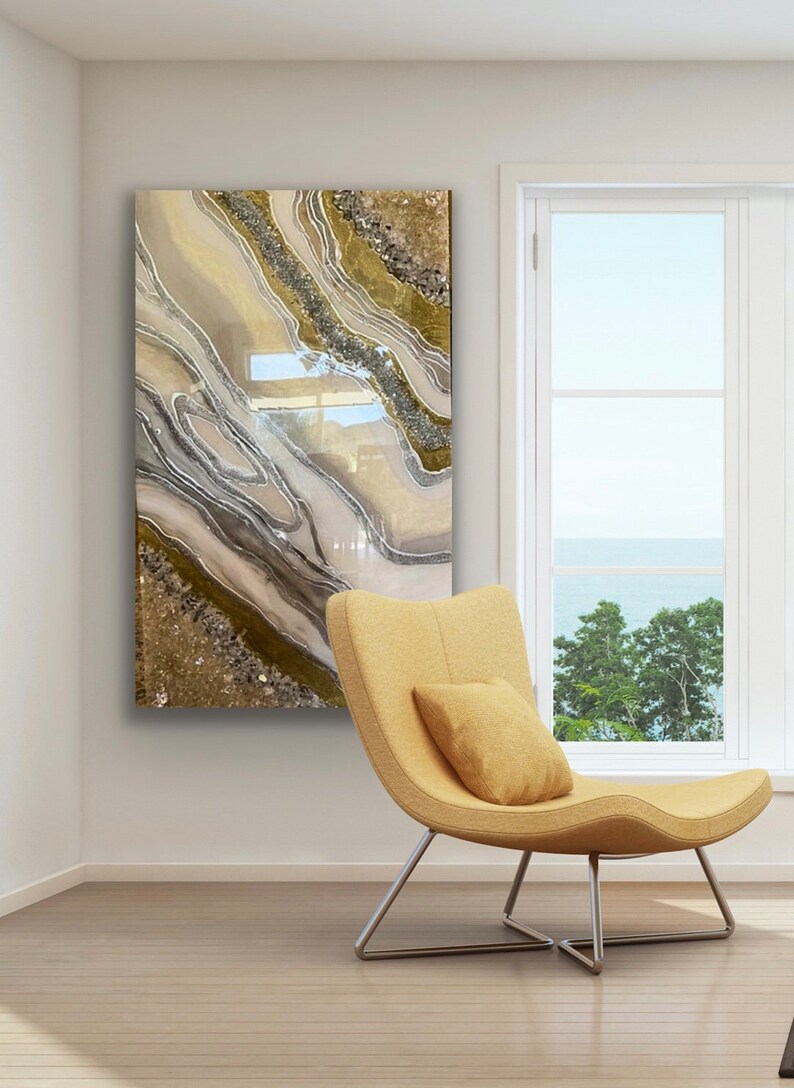 Puede incluir: Una pintura abstracta contempor&aacute;nea con l&iacute;neas de brillo dorado, marr&oacute;n y plateado que se arremolinan sobre un fondo blanco. La pintura est&aacute; colgada en una pared blanca en una habitaci&oacute;n con una silla amarilla y una ventana con vista al mar.