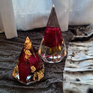 Op de afbeelding: Twee doorzichtige glazen ornamenten in de vorm van een piramide met gedroogde rode rozen en bladgoud. De ornamenten liggen op een grijs stoffen oppervlak.