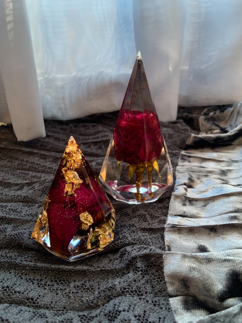 Op de afbeelding: Twee doorzichtige glazen ornamenten in de vorm van een piramide met gedroogde rode rozen en bladgoud. De ornamenten liggen op een grijs stoffen oppervlak.