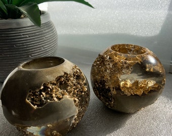 Op de afbeelding: Twee ronde, bruine kaarshouders met gouden vlokken. De kaarshouders zijn gemaakt van hars en hebben een getextureerd oppervlak.