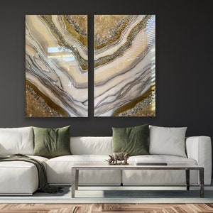 Puede incluir: Dos pinturas abstractas con un esquema de color blanco, gris y dorado. Las pinturas est&aacute;n en una pared gris oscuro sobre un sof&aacute; blanco con almohadas de tiro verdes.