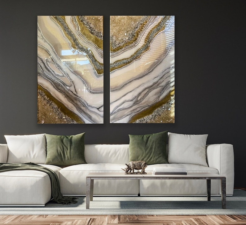 Puede incluir: Dos pinturas abstractas con un esquema de color blanco, gris y dorado. Las pinturas est&aacute;n en una pared gris oscuro sobre un sof&aacute; blanco con almohadas de tiro verdes.