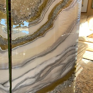 Puede incluir: Dos grandes paneles de arte rectangulares de resina blanca con purpurina dorada y plateada y cristales de cuarzo transparentes incrustados en la resina. Los paneles tienen un patr&oacute;n de remolino, similar al del &aacute;gata.