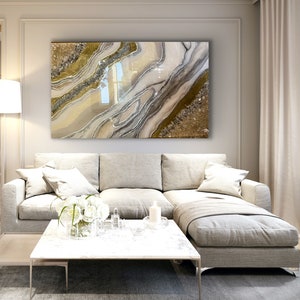 Puede incluir: Un sof&aacute; seccional blanco con una manta de tiro gris claro y una mesa de caf&eacute; blanca con un jarr&oacute;n de flores blancas. Una gran pintura abstracta con colores dorado, plateado y blanco cuelga de la pared detr&aacute;s del sof&aacute;.
