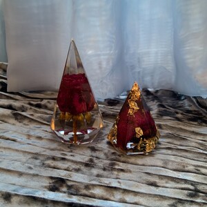 Op de afbeelding: Twee doorzichtige glazen ornamenten in piramidevorm met een rode roos erin. Een ornament heeft gouden bladaccenten.