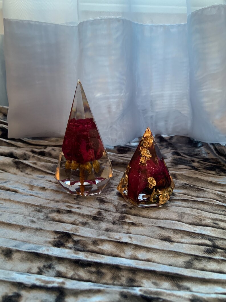 Op de afbeelding: Twee doorzichtige glazen ornamenten in piramidevorm met een rode roos erin. Een ornament heeft gouden bladaccenten.