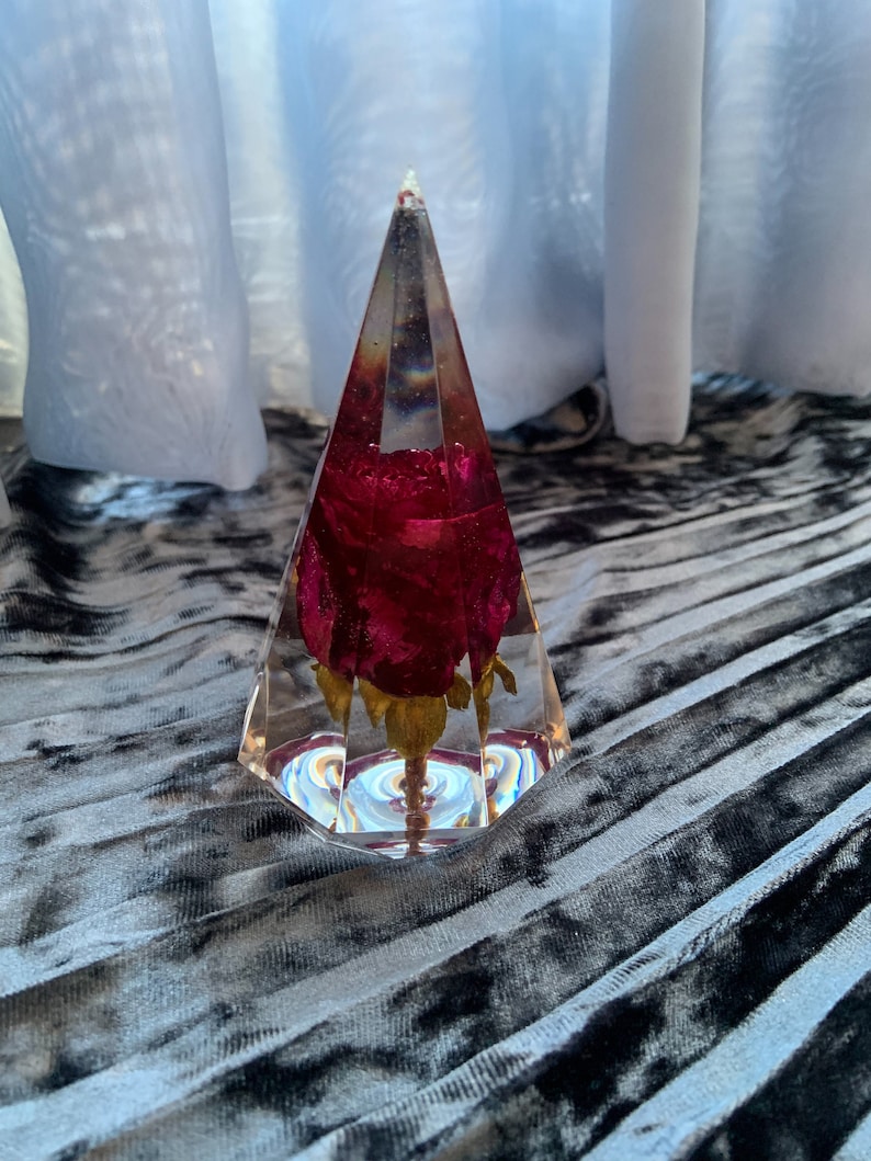Op de afbeelding: Een heldere glazen piramide met een geconserveerde rode roos erin. De piramide staat op een grijs en zwart stoffen oppervlak.