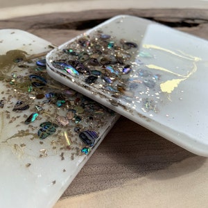 Könnte beinhalten: Zwei weiße quadratische Untersetzer mit schimmernden und goldenen Glitzer, die in klarem Harz eingebettet sind. Die Untersetzer sind übereinander gestapelt.