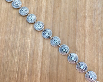 Könnte beinhalten: Silberarmband mit einer Kette und runden, dekorativen Anhängern. Jeder Anhänger hat ein erhabenes, florales Design und einen klaren Strassstein in der Mitte.